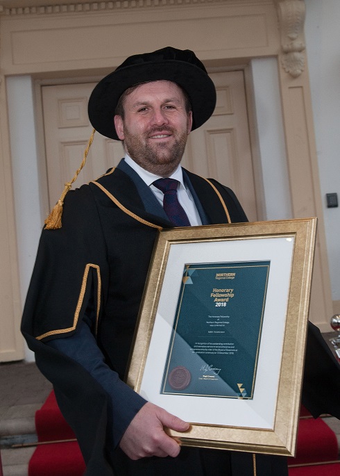 Male honorary fellow in his gown holding a framed certificate outside Galgorm Resort and Spa