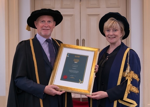 Female in graduation robe presenting a framed certificate to a male in graduation robe