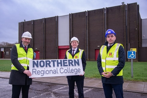Three males wearing high vis vests and hard hats holding College logo board